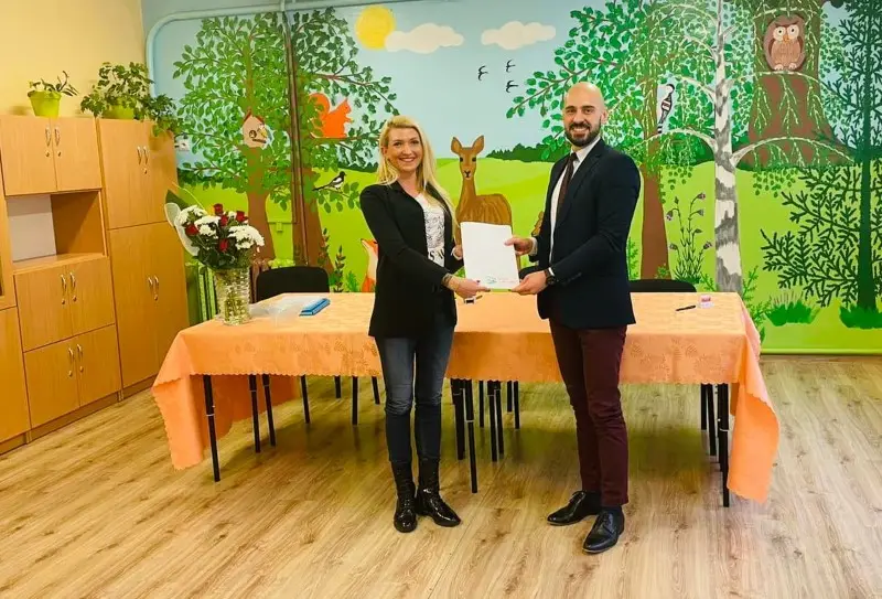 Zdjęcie w orientacji poziomej przedstawia kobietę i mężczyznę stojących w sali przedszkolnej, trzymających wspólnie biały dokument. Kobieta po lewej stronie ubrana jest w czarny żakiet, jeansy i wysokie czarne buty, podczas gdy mężczyzna po prawej ma na sobie elegancką marynarkę, białą koszulę z krawatem oraz ciemne spodnie. Za nimi znajduje się stół przykryty pomarańczowym obrusem, na którym stoi wazon z kwiatami, oraz kolorowy mural na ścianie przedstawiający leśne zwierzęta i drzewa. Jasne drewniane szafki wkomponowane są w zabudowę po lewej stronie, a podłoga wykonana jest z jasnych paneli imitujących drewno. Sceneria sugeruje oficjalne spotkanie w edukacyjnym otoczeniu, utrzymane w przyjaznym, szkolnym klimacie. Całość kadru jest dobrze oświetlona, co podkreśla profesjonalizm osób oraz bajkowy charakter wnętrza sali.