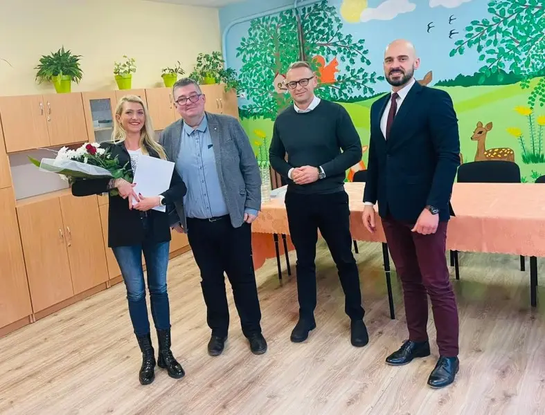 Zdjęcie w orientacji pionowej przedstawia grupę czterech osób, jedną kobietę i trzech mężczyzn, stojących w jasnej sali przedszkolnej lub szkolnej. Kobieta po lewej stronie trzyma bukiet kwiatów oraz białą kopertę, ubrana jest w czarną marynarkę, dżinsy i czarne botki. Obok niej stoi mężczyzna w szarej marynarce, a po prawej stronie widać dwóch kolejnych mężczyzn – jednego w ciemnym swetrze i drugiego w eleganckiej marynarce z bordowymi spodniami. Tło stanowi drewniana zabudowa meblowa oraz kolorowy mural na ścianie przedstawiający leśne zwierzęta, takie jak sarenka i wiewiórka, oraz drzewa. Na pierwszym planie widoczny jest stół przykryty brzoskwiniowym obrusem, a całe wnętrze utrzymane jest w ciepłej, przyjaznej kolorystyce z jasną podłogą imitującą drewno.