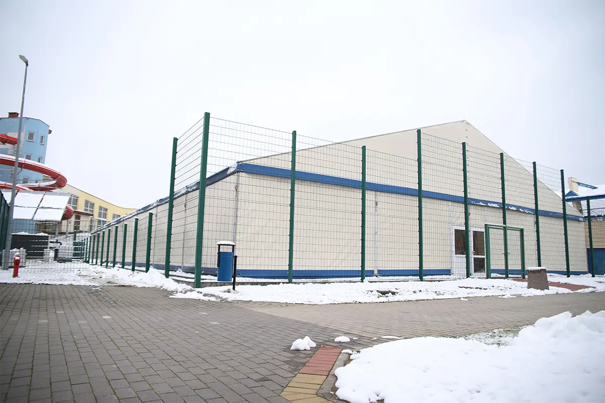 Zdjęcie przedstawia dużą halę namiotową o jasnych ścianach i dwuspadowym dachu, ogrodzoną wysokim zielonym płotem z siatki stalowej. Obiekt o proporcjach boków 3:2 został uchwycony w zimowej scenerii, gdzie zalegający śnieg przykrywa wybrukowany chodnik prowadzący w stronę wejścia do hali. Po lewej stronie kadru widoczna jest część konstrukcji zjeżdżalni wodnej oraz nowoczesny budynek w tle, co sugeruje lokalizację na terenie kompleksu rekreacyjnego. Całość utrzymana jest w chłodnej, stonowanej kolorystyce, podkreślającej pochmurny, zimowy dzień. Charakterystycznym elementem na pierwszym planie jest niebieski kosz na śmieci umieszczony bezpośrednio przy ogrodzeniu terenu.